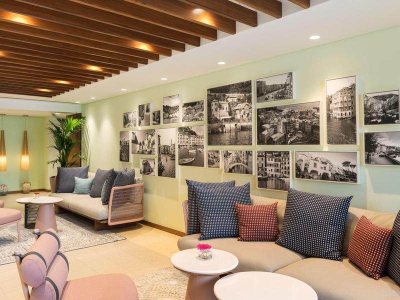 Moderne hotel lounge met crèmesofa's, donkere kussens, ronde tafels en zwart-witte foto aan de muur.