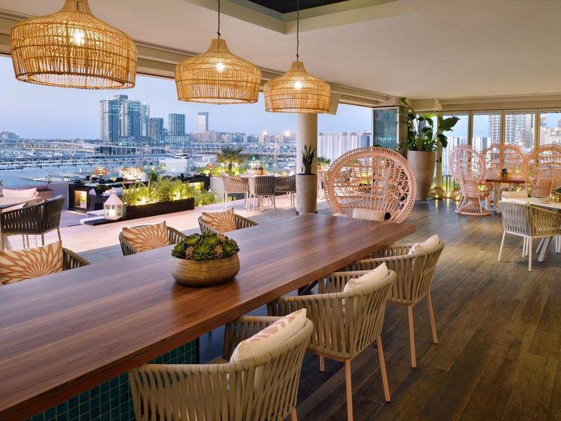 Gezellig buitenterras met houten tafels, rieten stoelen, hanglampen en uitzicht op de stad.