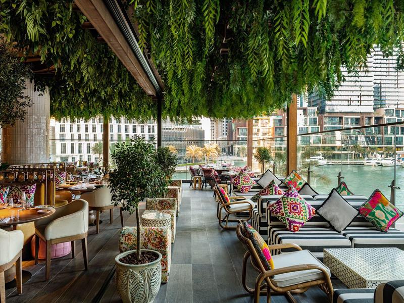 Buitengebied restaurant met tafels, stoelen, kussens, planten en uitzicht op de stad.