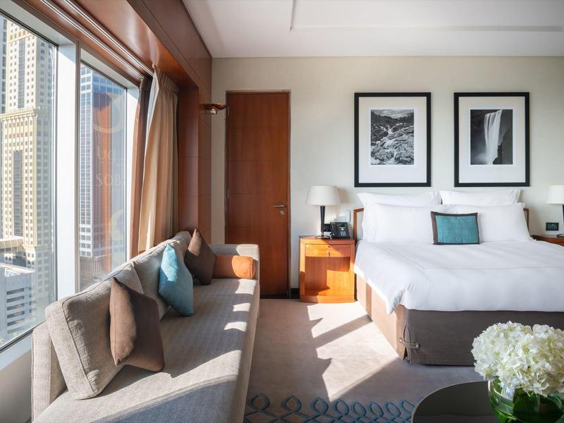 Bright hotel room with large window, double bed, sofa, and city view.