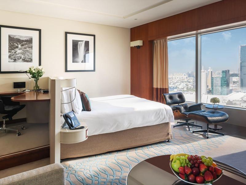 Modern hotel room with bed, desk, comfortable chairs, and large window overlooking city skyline.