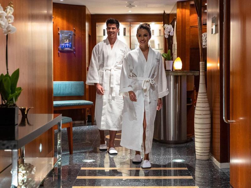 Two people in white robes walk through a stylish spa lobby with warm wood decor.