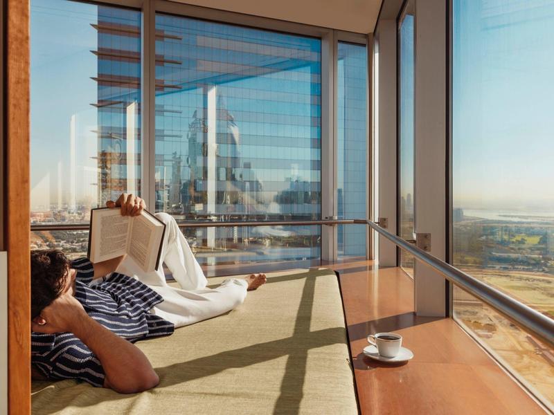 Person relaxing and reading in a room with large windows overlooking a city.