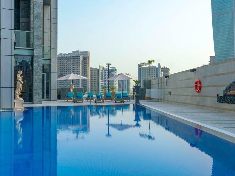 Blue hotel rooftop pool with city view and sun umbrellas under clear sky.