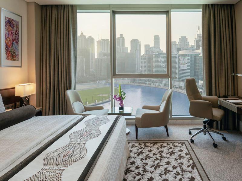 Modern hotel room with two beds, armchair, desk, and city view.