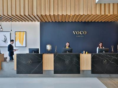 Moderne Hotelrezeption mit drei schwarzen Empfangstheken und Personal vor dunkler Wand mit Goldschriftzug VOCO.