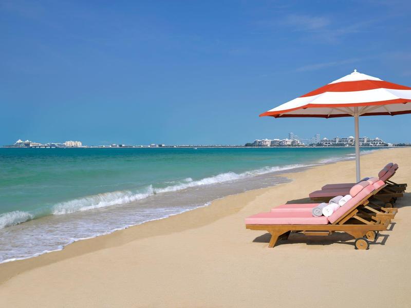Leeg strand met twee ligstoelen en parasol bij kalme zee onder blauwe lucht.