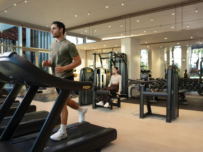 Een man loopt op een loopband in een lichte fitnessruimte met spiegels en trainingsapparatuur.