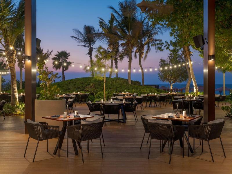 Avondterras van een restaurant aan zee met palmbomen, sfeerverlichting en gedekte tafels.