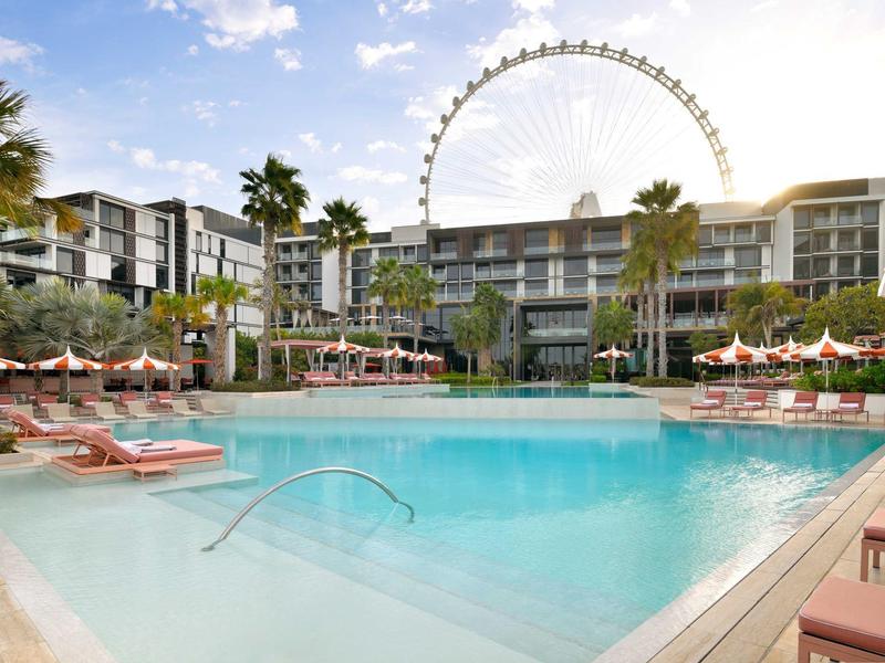 Modern hotelzwembad met ligstoelen en reuzenrad op de achtergrond bij zonnig weer.