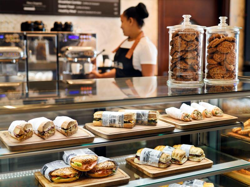 Vetrina con panini confezionati e biscotti in barattoli, barista al lavoro alla macchina per il caffè sullo sfondo.