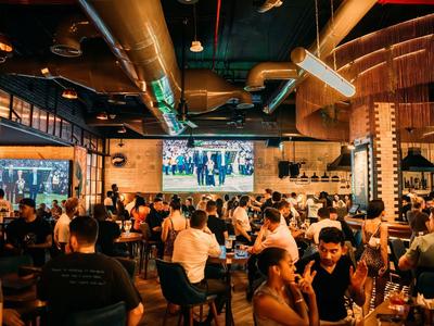 Druk restaurant met gasten, moderne lampen en een grote tv aan de muur.
