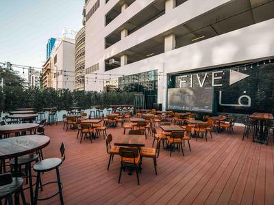 Venkovní jídelní prostor s dřevěnou terasou a stolky a židlemi před moderní budovou.