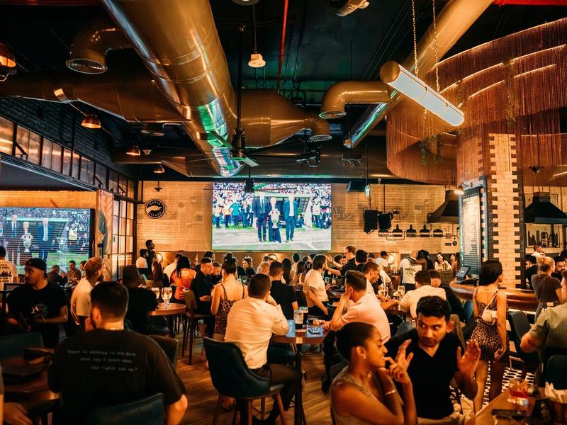 Druk restaurant met gasten, moderne lampen en een grote tv aan de muur.
