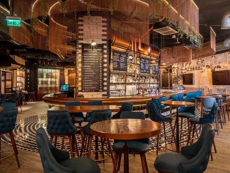 Moderne hotelbar met ronde houten tafels en blauwe fluwelen stoelen in warm verlichte ruimte.