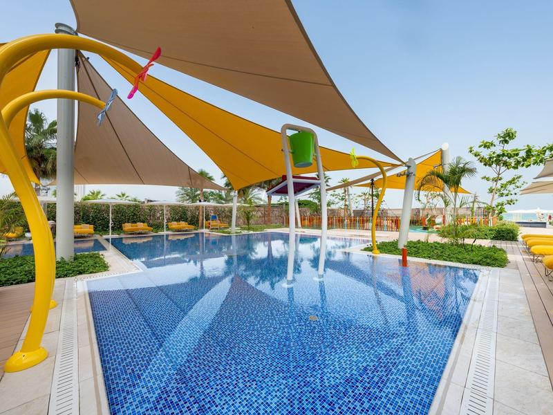 Modern outdoor pool with sun sails and water features in a hotel garden.