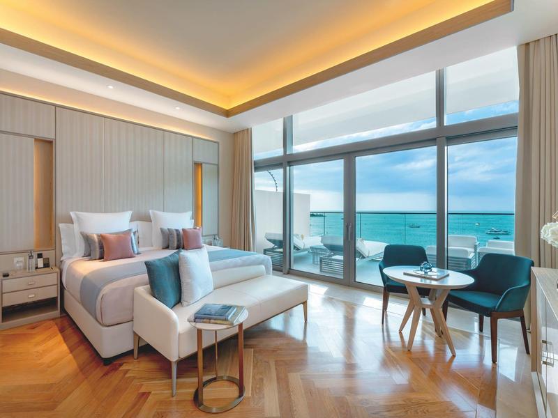 Modern hotel room with large bed, seating area, and panoramic windows overlooking the sea.