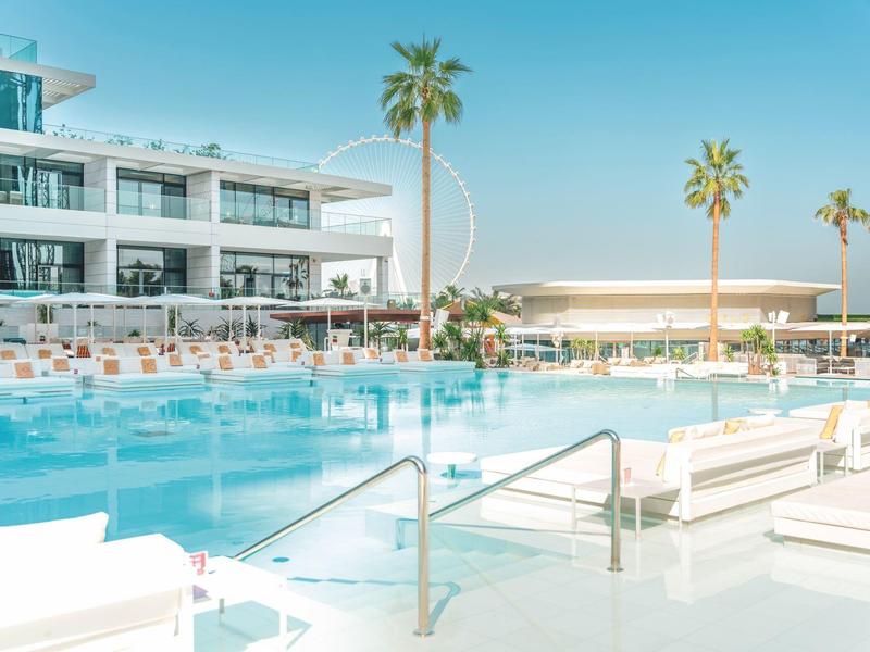 Modern outdoor pool with loungers and palm trees in front of a hotel building under clear sky.