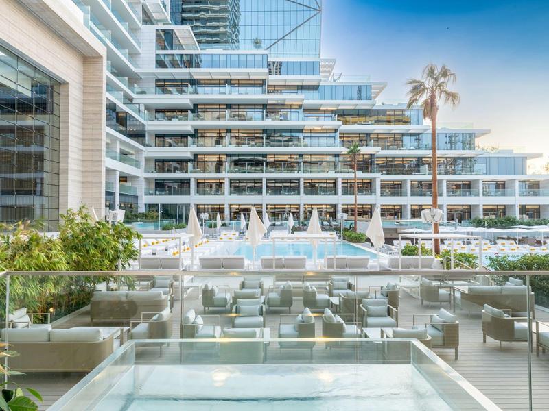Modern hotel pool area with glass railing, lounge chairs, and tall building facade in the background.