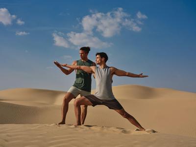Zwei Männer praktizieren Yoga im Sand der Dünen unter blauem Himmel.