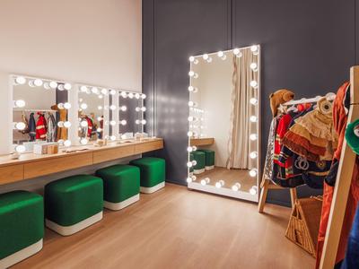 Šatna s osvětlenými zrcadly, zelenými stoličkami a barevnými kostýmy na věšácích.