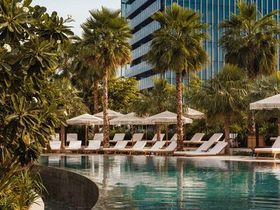 Moderner Hotelpool mit Liegestühlen, Sonnenschirmen und Palmen vor Hochhausfassade.