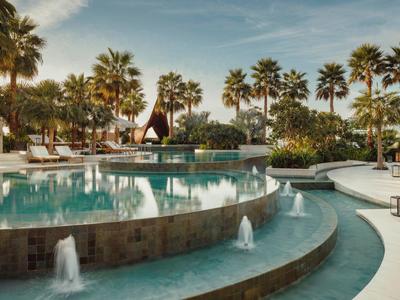 Großer Pool mit Wasserfontänen und Palmen an einem sonnigen Tag in einem Hotelgarten.