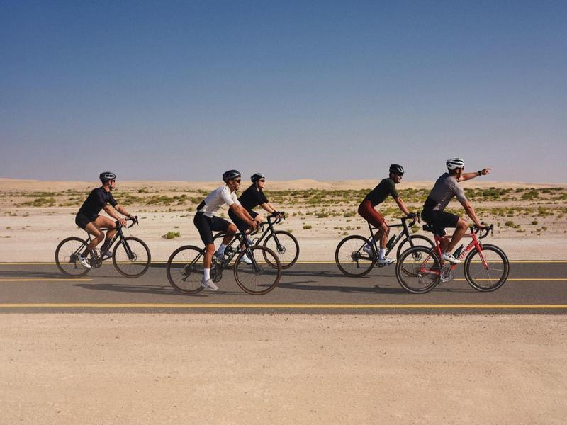 Five cyclists ride side by side on a road beside a desert under a clear sky.