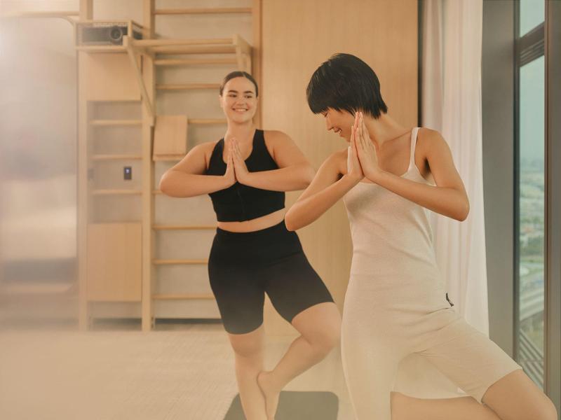 Two women practicing yoga in a bright room with large windows.