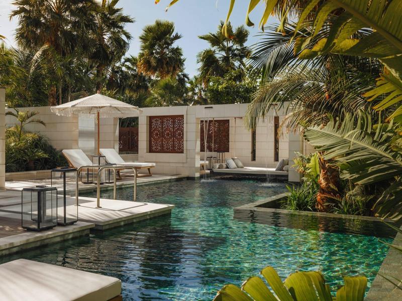 Pool with lounge chairs and umbrellas in front of a house, surrounded by palm trees.