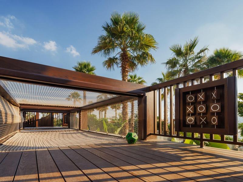 Modern wooden pavilion with sunshade and tropical palm trees under blue sky.