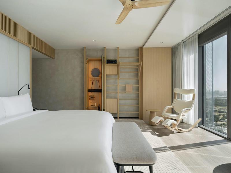 Modern hotel room with large bed, balcony, and city view in daylight.