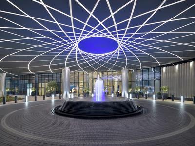 Moderner Zugang mit rundem Brunnen und leuchtendem, blauem Licht unter einem geometrischen Gitterdach.