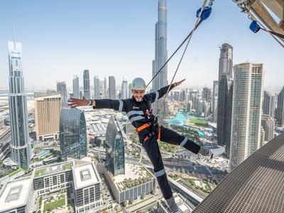 Person im Klettergurt schwebt an Seil vor Wolkenkratzern in moderner Stadt bei klarem Himmel.