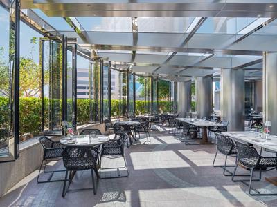 Moderner verglaster Außenbereich mit schwarzen Stühlen und Tischen, viel Tageslicht und grünem Sichtschutz.