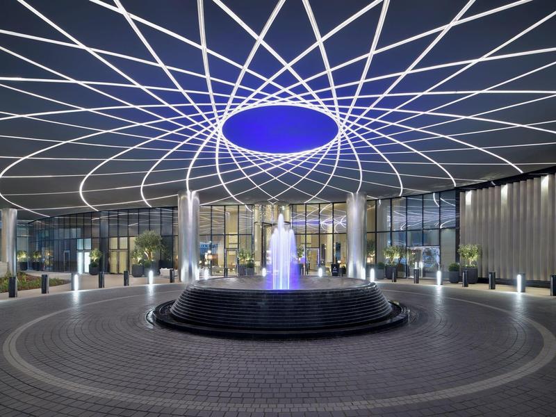 Moderner Zugang mit rundem Brunnen und leuchtendem, blauem Licht unter einem geometrischen Gitterdach.