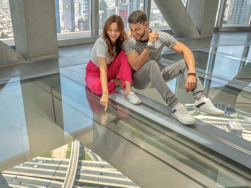 Zwei Personen sitzen auf einem Glasboden mit Blick auf eine Stadt und Hochstraßen darunter.