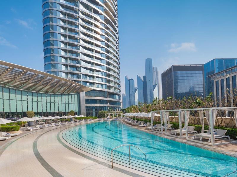 Großer Außenpool mit runden Stufen, Liegen in Reihe und moderner Stadt im Hintergrund bei Sonnenschein.