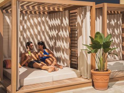 Zwei Frauen entspannen auf einer großen Sonnenliege unter einem Holzpavillon mit Sonnenschein und Pflanzen.