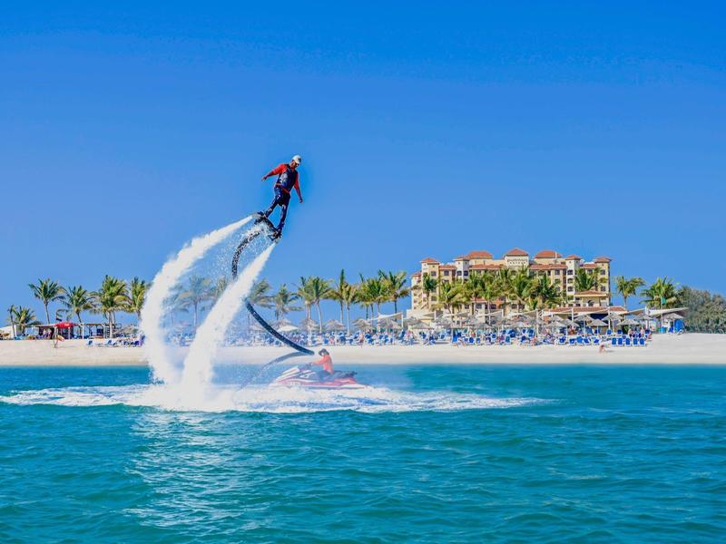 Person mit Flyboard über blauem Wasser vor Insel mit Hotel und Strand