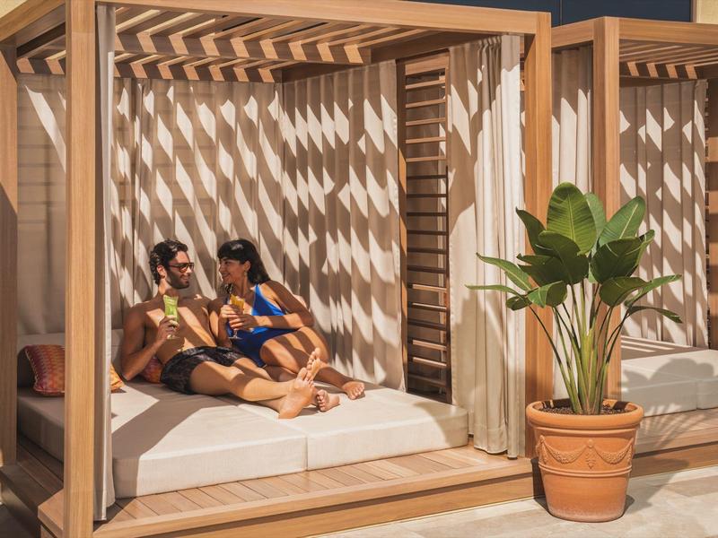 Zwei Frauen entspannen auf einem Tagesbett mit Holzrahmen an einem sonnigen Poolbereich.