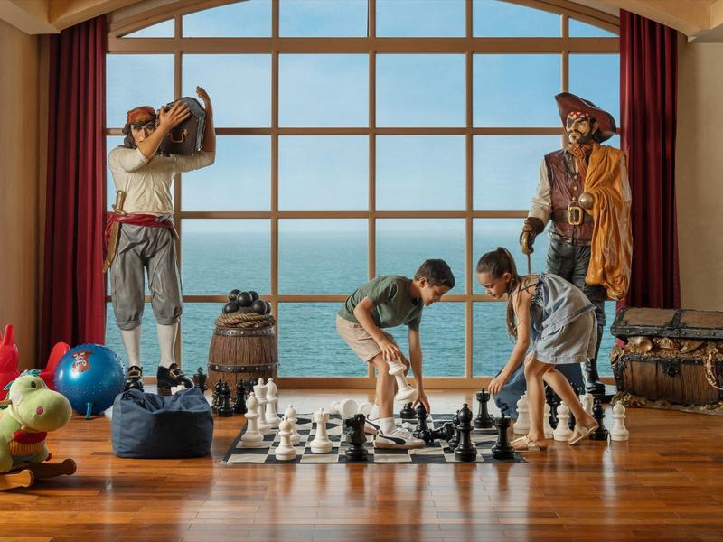 Drei Personen spielen Schach vor einem Fenster mit Meerblick in einem Holzraum.