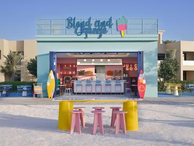 Colorful beach bar with seating and ice cream counter in front of buildings under blue sky.