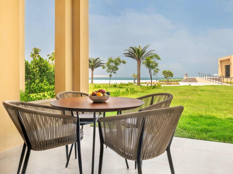 Terrasse avec table et chaises donnant sur une pelouse verte et des palmiers près de la plage.