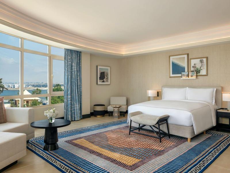 Chambre d'hôtel lumineuse avec grand lit, tapis, fauteuils et vue sur la ville.