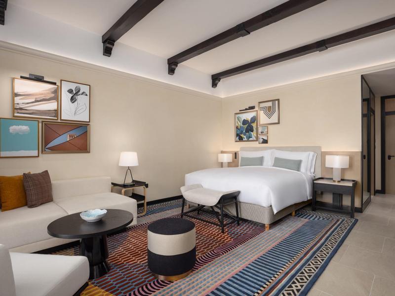 Chambre d'hôtel lumineuse avec lit double, canapé, tapis coloré et tableaux muraux sous poutres apparentes