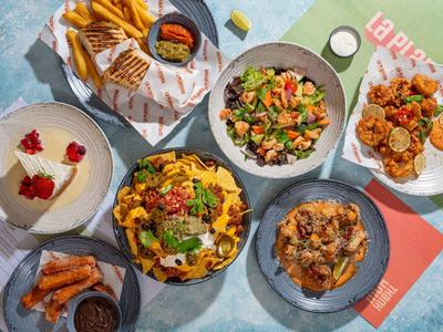 Verschiedene bunte Gerichte mit Pommes, Burgern, Salat, Nachos und Dessert auf einem Tisch.