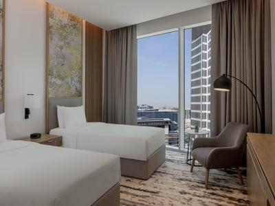 Helles Hotelzimmer mit zwei Einzelbetten, grauem Sessel, großem Fenster und Blick auf Hochhäuser.