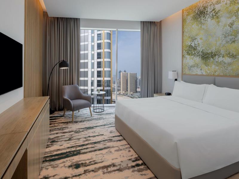 Modernes Hotelzimmer mit großem Bett, Teppichboden, Sessel und Stadtblick durch Panoramafenster.