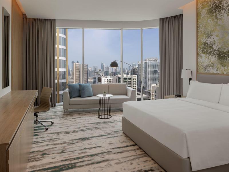 Helles Hotelzimmer mit großem Bett, Sofa, Schreibtisch und Panoramafenster mit Stadtblick.